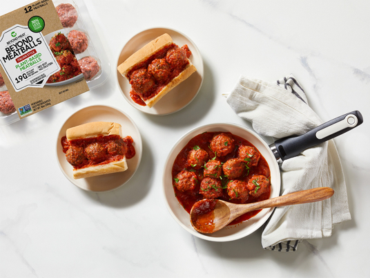 A white counter with a skillet of meatballs and tomato sauce sitting next two two meatball subs and a package of Beyond Meatballs