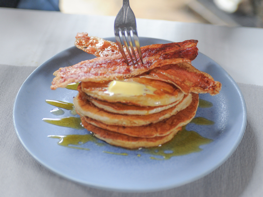 Higher Steaks lab grown bacon served on a stack of pancakes. The plate is drizzled with syrup and there's a melty pat of butter on the pancakes, as well. Someone is digging into the stack with a fork.