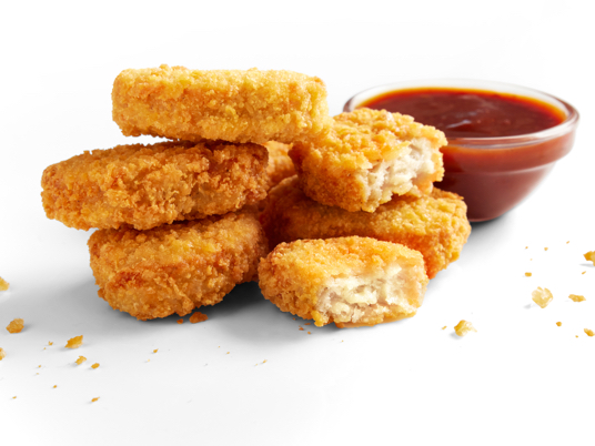 A stack of Imposible Chicken Nuggets. One of them is torn in half. There is a small bowl of sauce next to the stack, and there are crumbs around the nuggets.