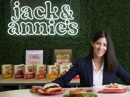 Photo of Annie Ryu sitting at a table. Above her, a fluorescent jack and annie's sign hangs on a plant wall.